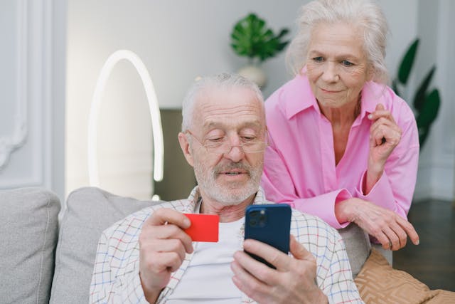 An older man holding his credit card and phone ready to make an online purchase as his wife looks from behind.